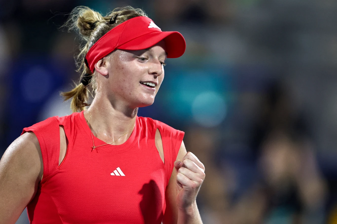 Tenisistka ubrana w czerwony strój i czerwoną czapkę z daszkiem, zaciśnięta pięść w geście triumfu, uśmiech na twarzy, rozmyte tło sugeruje dynamiczną atmosferę sportową.