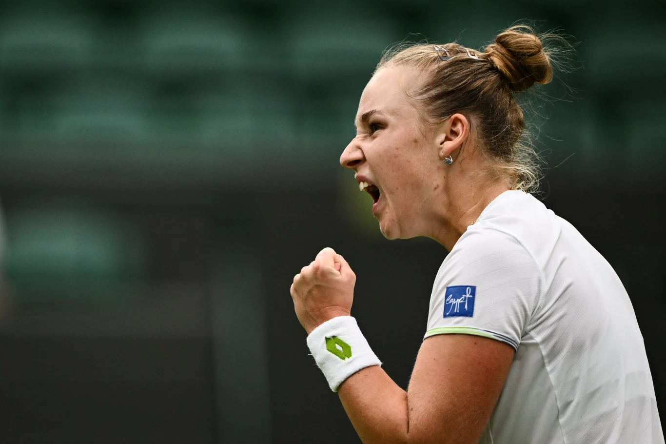 Zawodniczka tenisowa w białym stroju sportowym zaciśnięta pięść i wyraz silnych emocji, wyrażający triumf lub determinację podczas meczu.