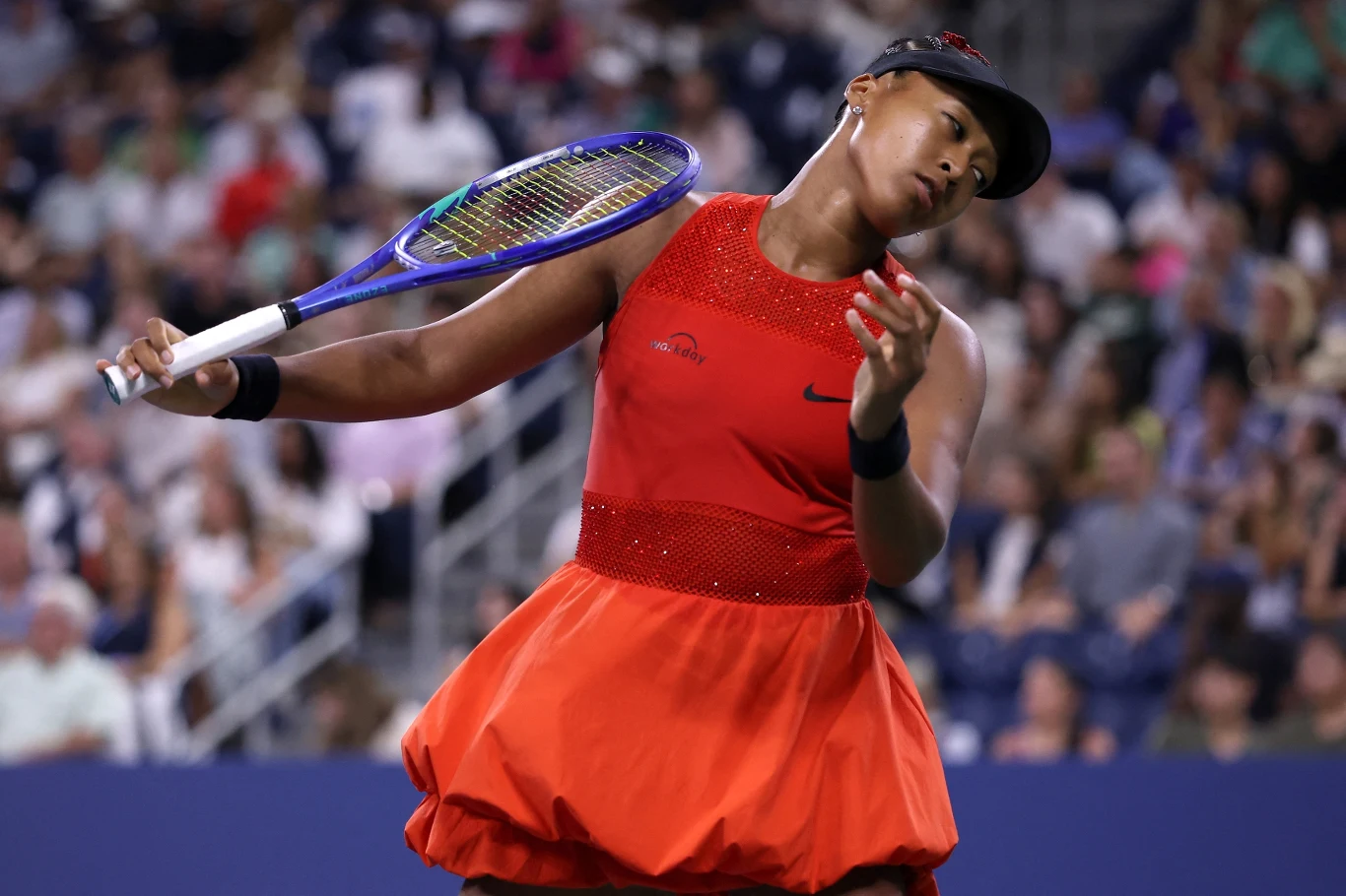 Naomi Osaka, US Open 2025 Tenisistka w czerwonej sukience wykonuje zamach rakietą podczas meczu na kortach, w tle rozmazana publiczność siedząca na trybunach.