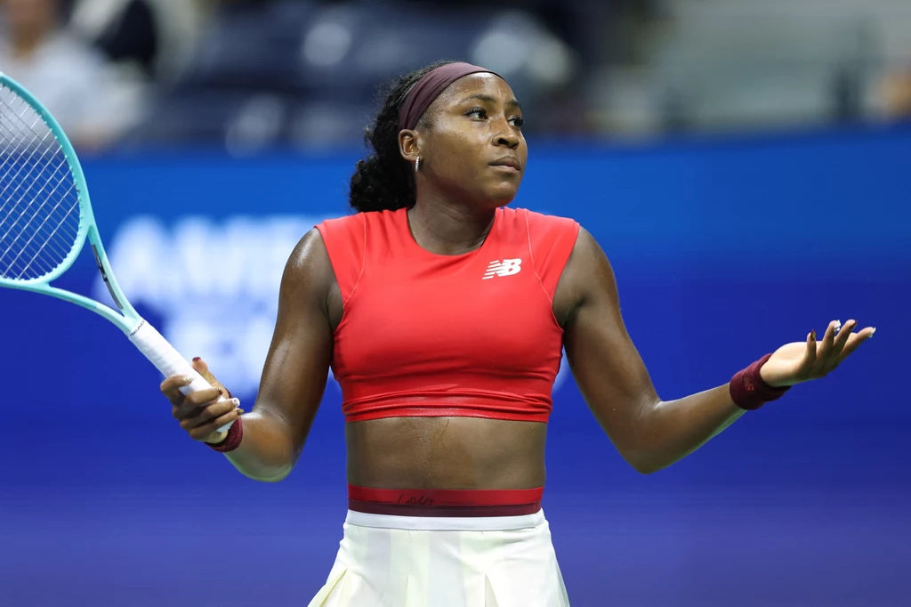Coco Gauff Zawodniczka tenisa ziemnego w czerwonym topie i białej spódnicy z rakietą w lewej dłoni wykonuje gest wzruszenia ramionami, stojąc na korcie podczas meczu.