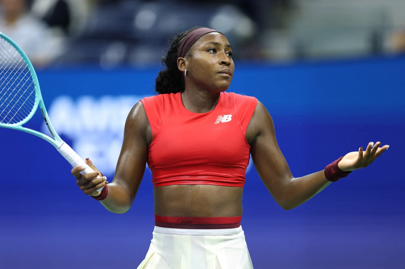 Zawodniczka tenisa ziemnego w czerwonym topie i białej spódnicy z rakietą w lewej dłoni wykonuje gest wzruszenia ramionami, stojąc na korcie podczas meczu.