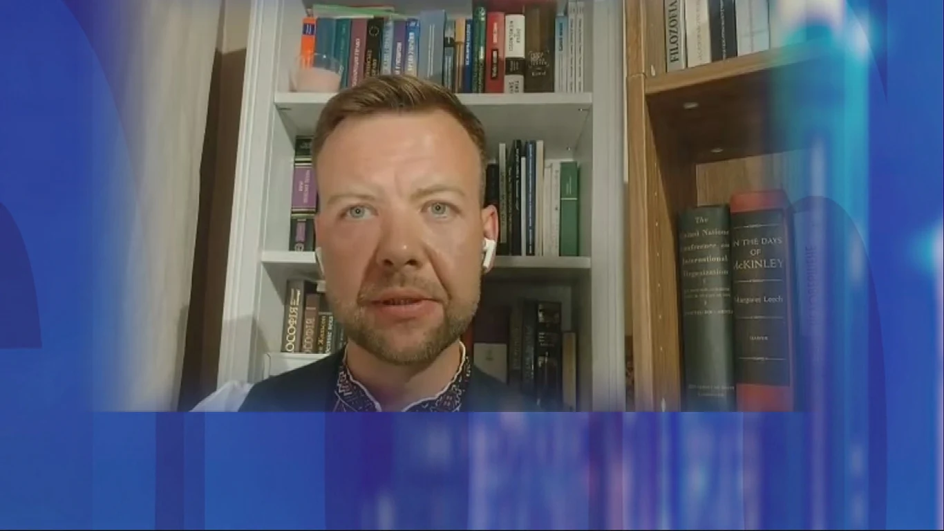 Dziennikarz Witalij Mazurenko Mężczyzna w średnim wieku słuchający przez douszną słuchawkę, siedzący na tle regału wypełnionego książkami, światło pada na jego twarz, po bokach widoczne są zniekształcone, niebieskie elementy graficzne.