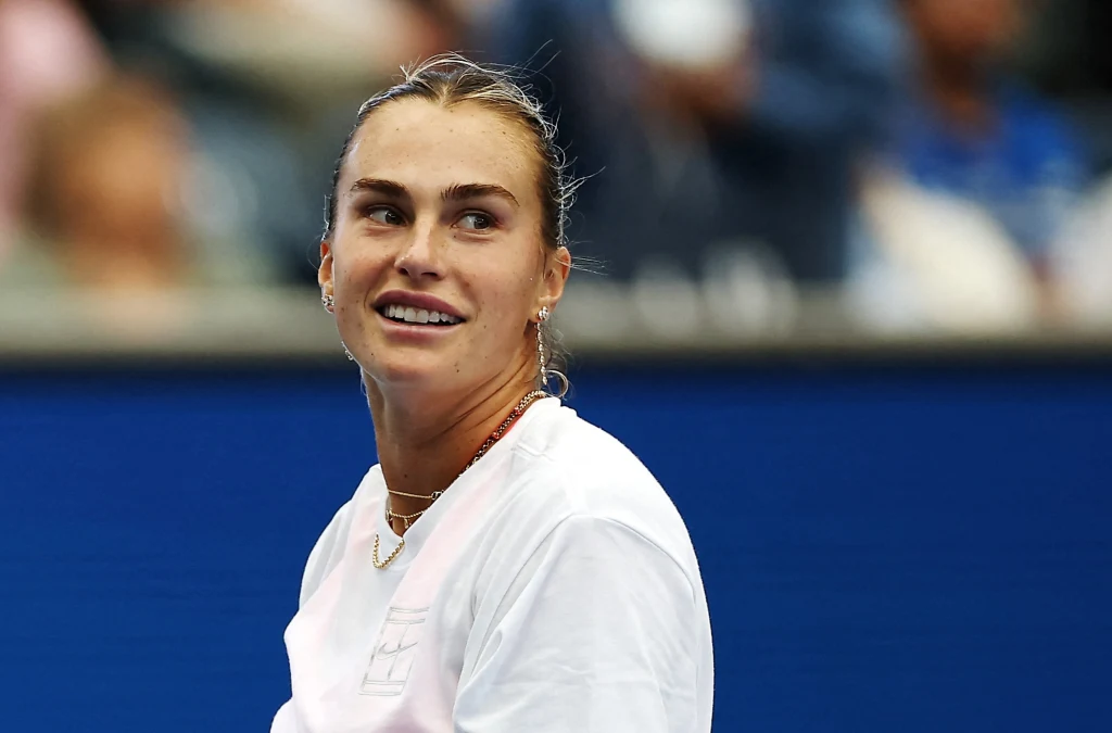 Aryna Sabalenka Uśmiechnięta młoda kobieta w białej koszulce patrząca w bok, tło zamazane i kontrastujące niebieskie barwy kortu tenisowego.