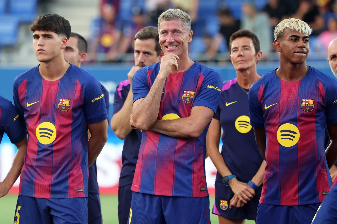 Pau Cubarsi, Robert Lewandowski i Lamine Yamal Grupa mężczyzn w klubowych koszulkach piłkarskich FC Barcelony stoi wspólnie na boisku, wszyscy uśmiechnięci i skupieni, część z nich ma ręce założone na piersi lub trzyma je przy twarzy, w tle widoczna jest trybuna stadionu.