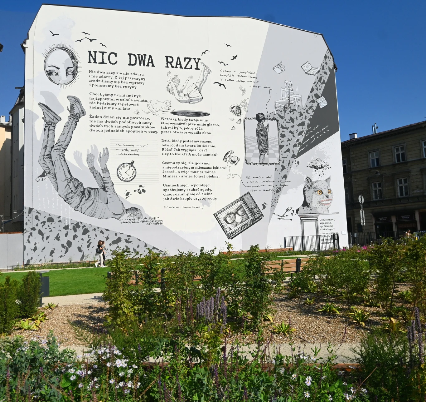 Duży mural na ścianie budynku z tekstem wiersza Wisławy Szymborskiej Nic Dwa Razy, ilustrowany licznymi czarno-białymi rysunkami przedstawiającymi postacie, przedmioty i symboliczne motywy, w tle widoczna zieleń miejskiego ogrodu.