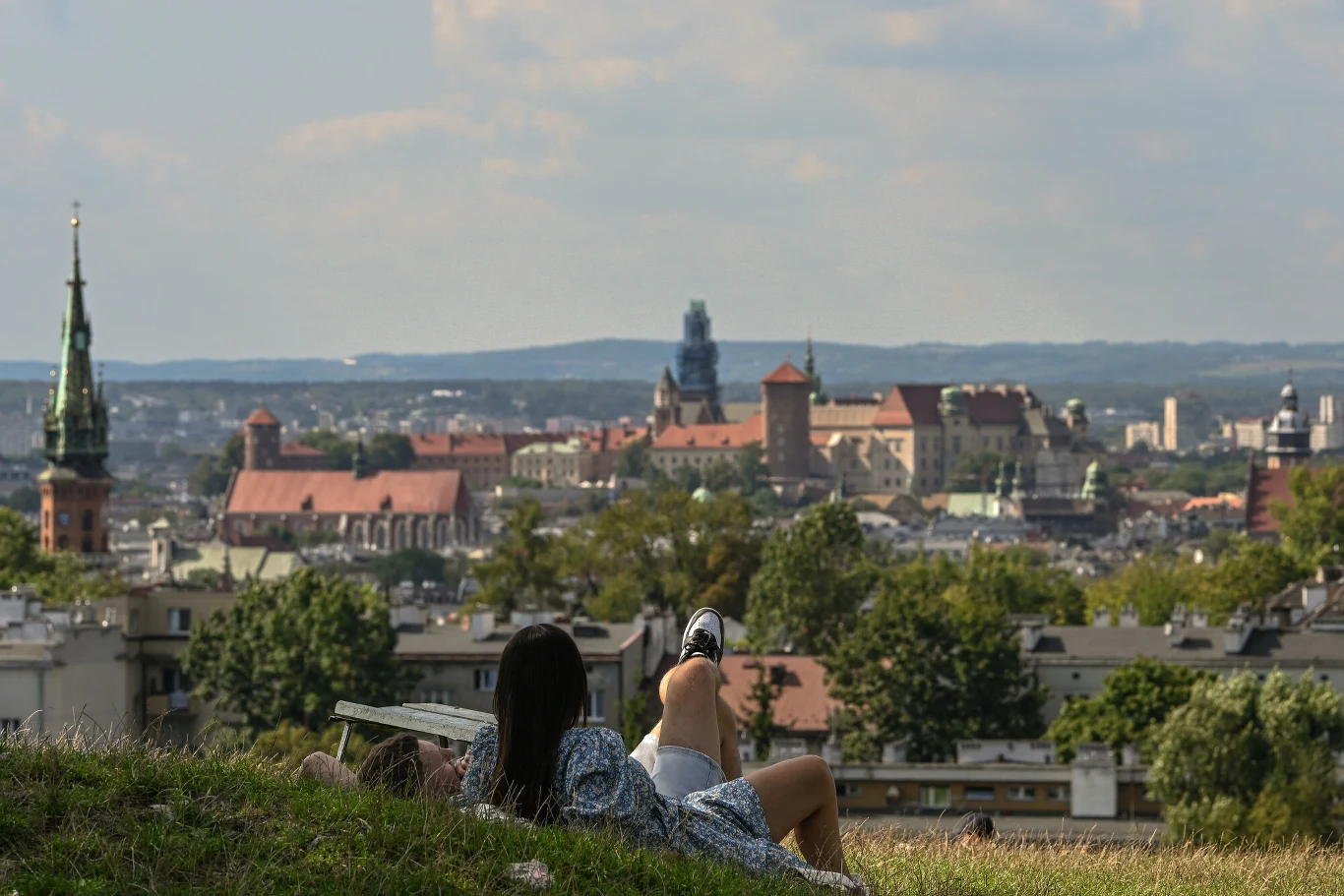 Osoba leżąca na trawie z widokiem na panoramę Krakowa, z wyraźnie widocznym Zamkiem Królewskim na Wawelu, otoczonym zielenią i zabudowaniami miejskimi, na pierwszym planie relaksująca się kobieta z książką.