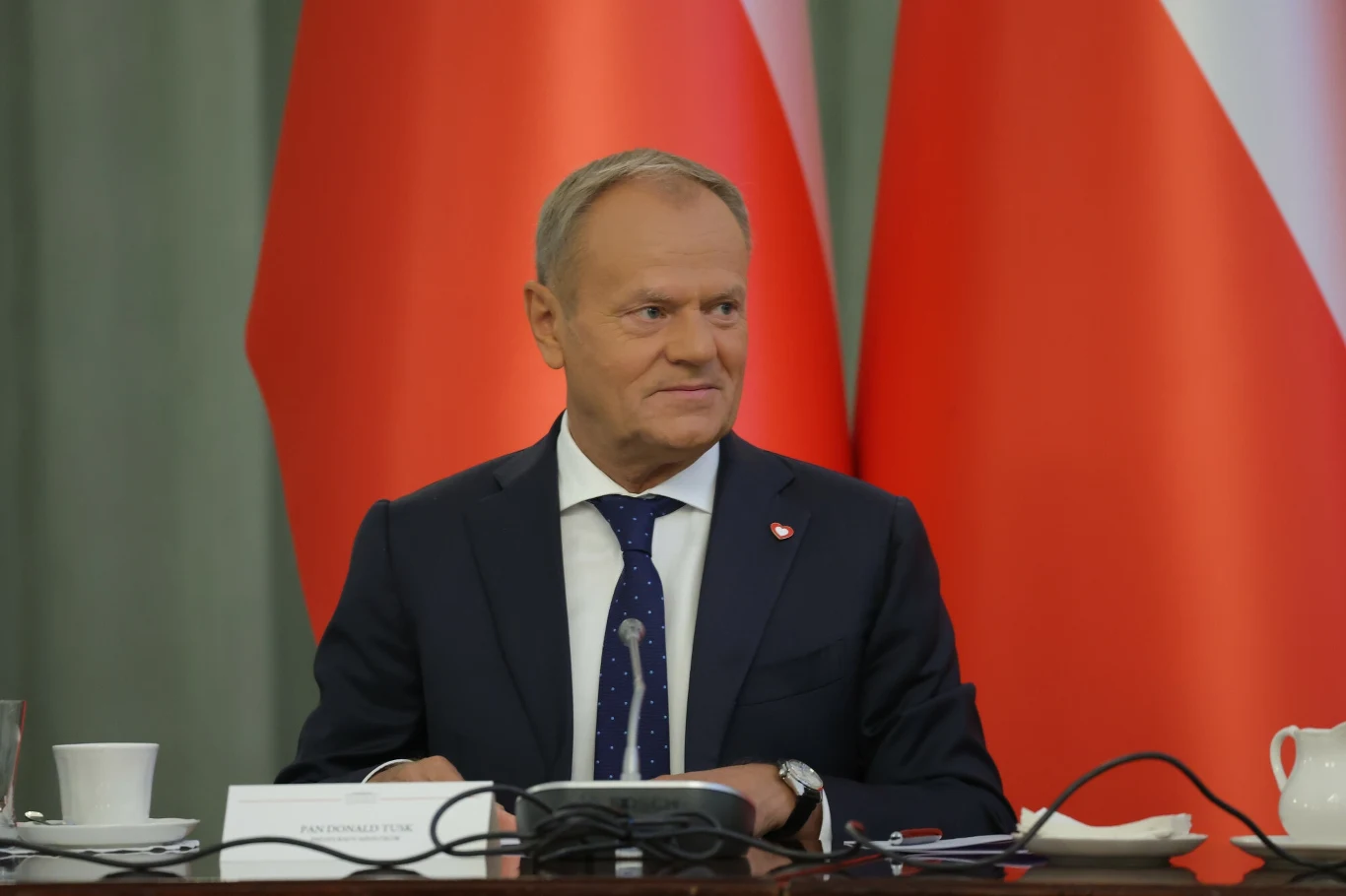 Donald Tusk zabrał głos przed niejawną częścią Rady Gabinetowej Mężczyzna w garniturze siedzący przy stole konferencyjnym, na tle polskich flag, z notatkami, filiżanką oraz mikrofonem przed sobą.