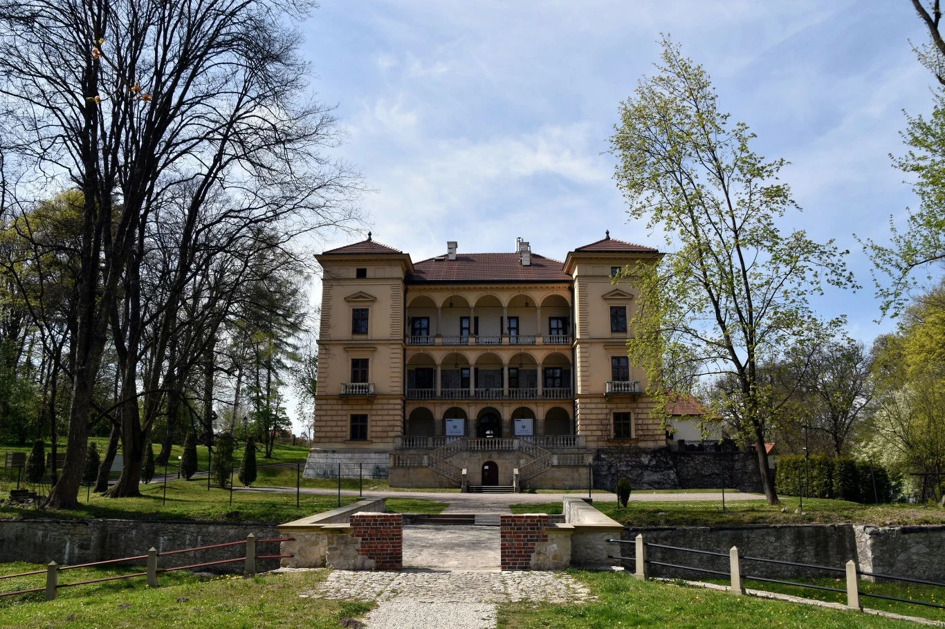 Renesansowy pałac z jasną fasadą z ozdobnymi balkonami położony w otoczeniu zieleni i starych drzew, prowadzi do niego szeroka ścieżka z niewielkim mostkiem nad suchą fosą.