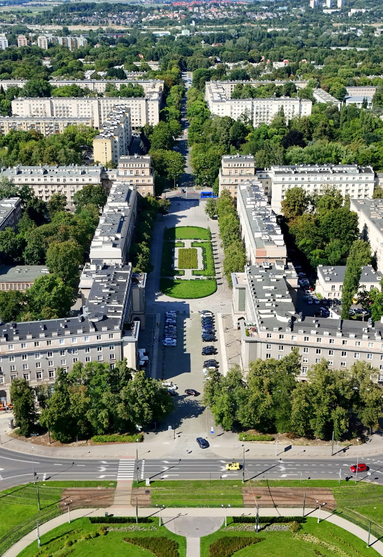 Nowa Huta. Widok na plac Centralny i al. Róż Panorama osiedla z charakterystyczną, zieloną aleją i symetrycznie ustawionymi budynkami mieszkalnymi otoczonymi drzewami, widoczne chodniki, ulice i zaparkowane samochody, w tle rozległy teren zielony.
