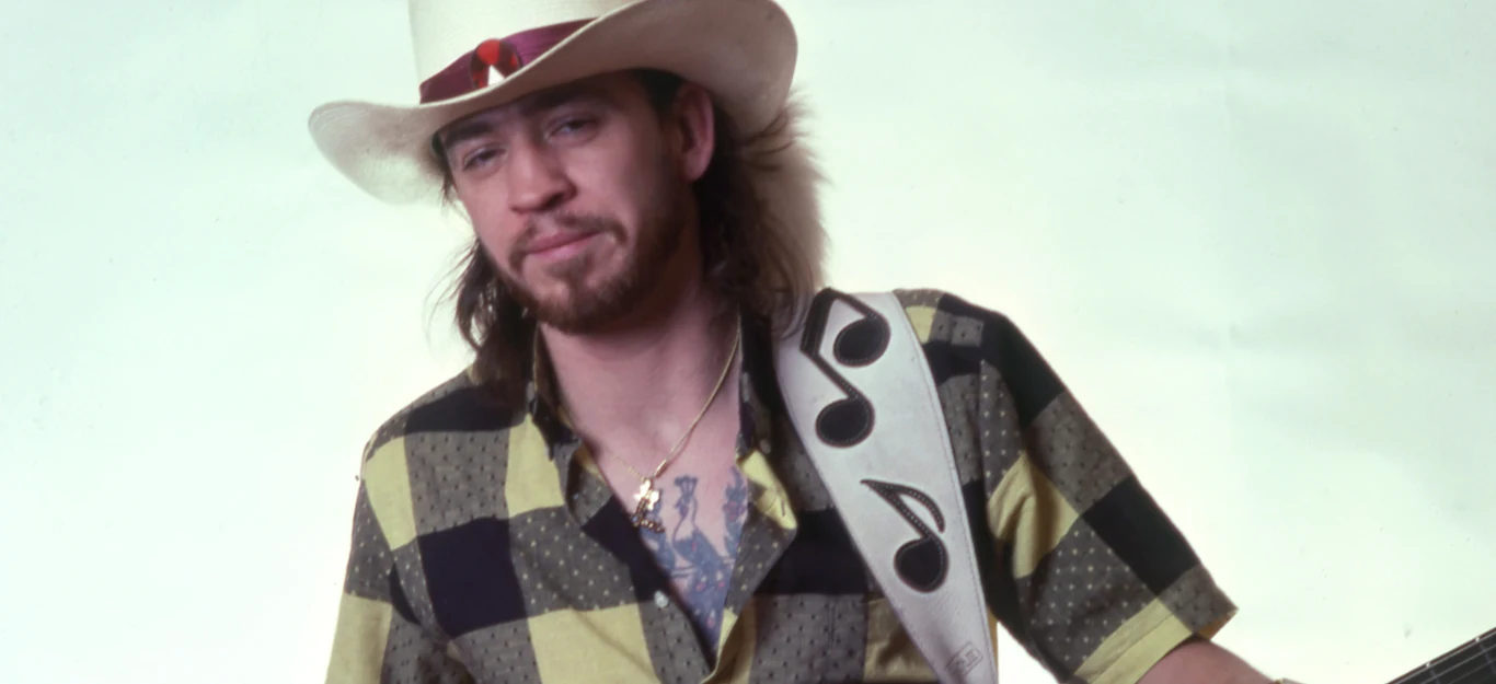 Stevie Ray Vaughan Mężczyzna w kapeluszu z szerokim rondem, ubrany w koszulę w dużą kratę, trzyma gitarę z charakterystycznym paskiem z motywem nut muzycznych, w tle jednolita jasna powierzchnia.