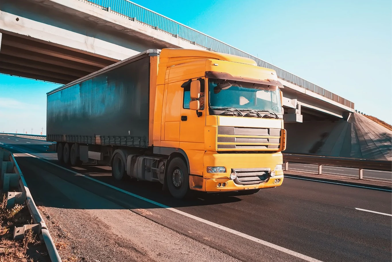 Okresowy zakaz jazdy nie dotyczy wszystkich pojazdów o DMC powyżej 12 ton. Żółta ciężarówka z dużą naczepą porusza się po asfaltowej drodze ekspresowej, w tle znajduje się betonowy wiadukt podświetlony promieniami słońca.