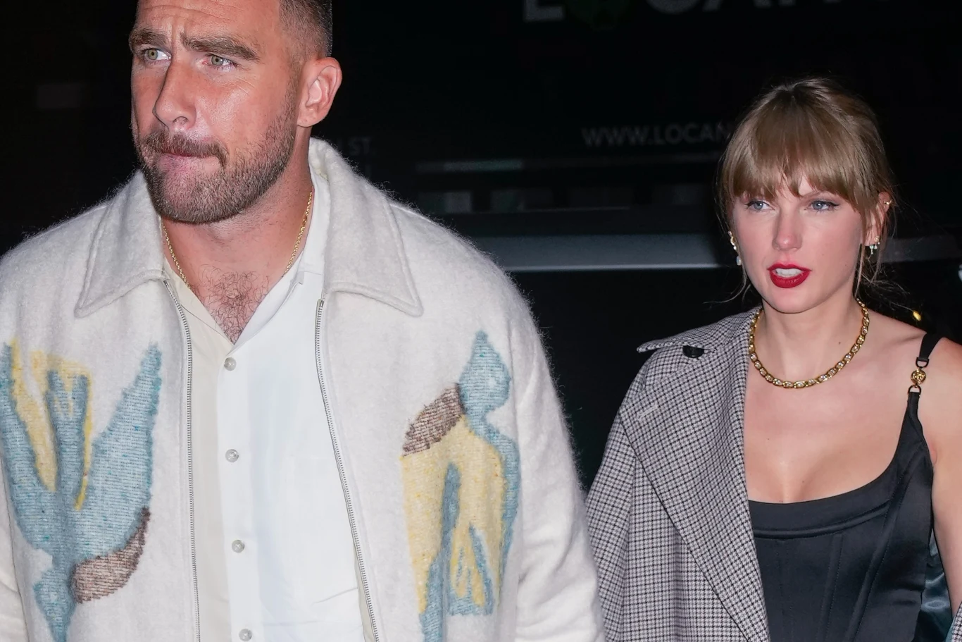 Travis Kelce i Taylor Swift są zaręczeni Mężczyzna w jasnej marynarce z wzorem idzie obok kobiety w czarnej sukience i płaszczu w kratę, oboje znajdują się na tle ciemnego otoczenia.