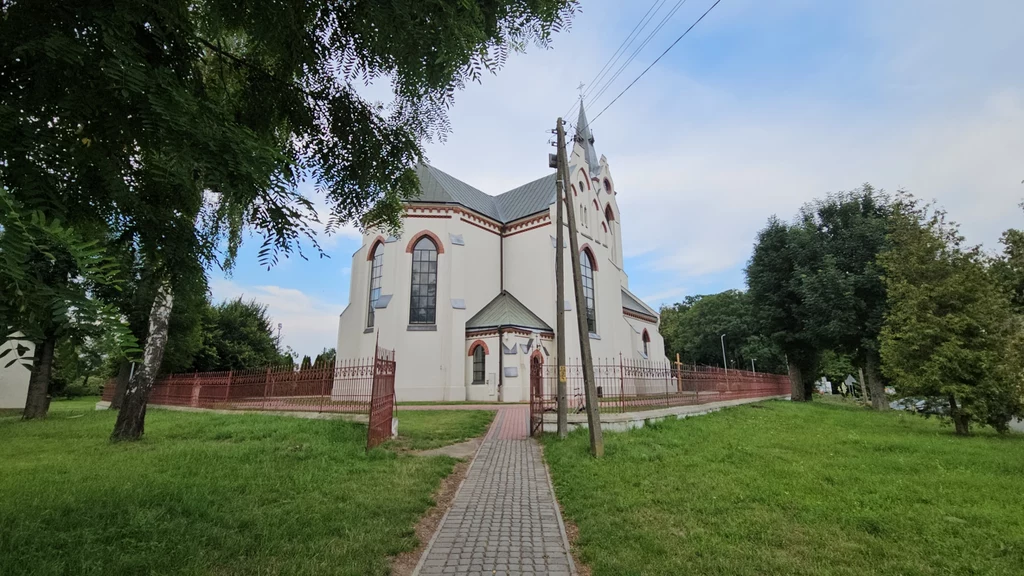 Kościół o jasnej elewacji z czerwonymi detalami architektonicznymi, otoczony ogrodzeniem i zielenią, z brukowaną ścieżką prowadzącą do głównego wejścia, w tle widoczne drzewa i błękitne niebo.