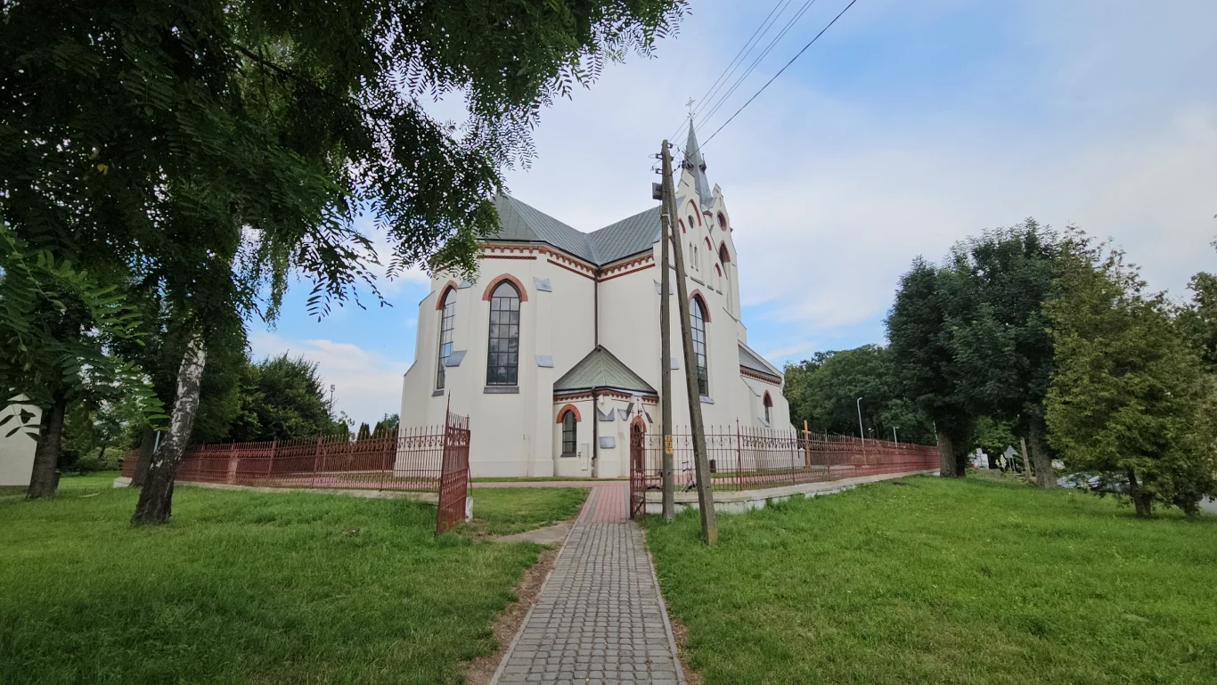 Kościół w Urzejowicach Kościół o jasnej elewacji z czerwonymi detalami architektonicznymi, otoczony ogrodzeniem i zielenią, z brukowaną ścieżką prowadzącą do głównego wejścia, w tle widoczne drzewa i błękitne niebo.