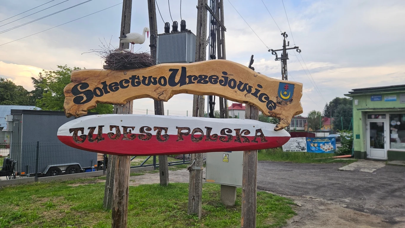 Urzejowice Drewniana tablica z napisem 'Sołectwo Urzejowice' oraz ozdobiona dużym gniazdem bociana i herbem, poniżej biało-czerwona deska z napisem 'Tu jest Polska', w tle zabudowania i słupy energetyczne.