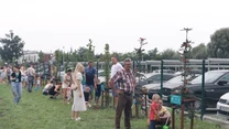 Grupa ludzi sadzi drzewa wzdłuż ogrodzenia przy parkingu, część osób pochyła się nad młodymi drzewkami, obok nich stoją dzieci z narzędziami ogrodniczymi, w tle zaparkowane samochody i zieleń miejskiego parku.