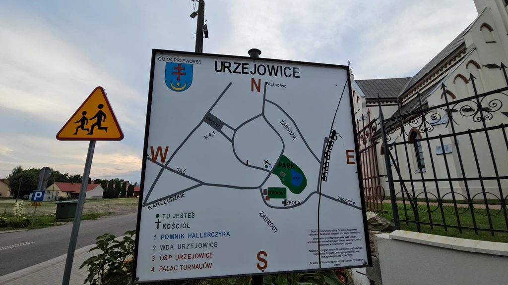 Tablica z mapą miejscowości Urzejowice z zaznaczonymi punktami orientacyjnymi, przy której widoczny jest znak drogowy ostrzegający o możliwym przejściu dzieci oraz fragment białego budynku z ogrodzeniem po prawej stronie.