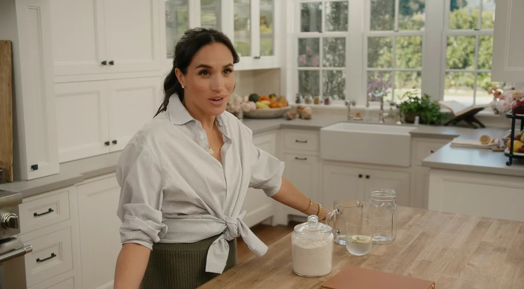 Meghan Markle zderzyła się z krytyką swojego programu Kobieta w jasnej koszuli stoi w jasnej, nowoczesnej kuchni, na drewnianym blacie przed nią stoi duży słoik z mąką, szklanka z wodą oraz notes; w tle widoczne miski z owocami i duże okna z widokiem na ogród.