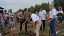 Grupa osób sadzi młode drzewka na polanie, używając łopat i sadzonek; wokół zgromadzeni są zarówno dorośli, jak i dzieci, a tło stanowią drzewa oraz otwarta przestrzeń.