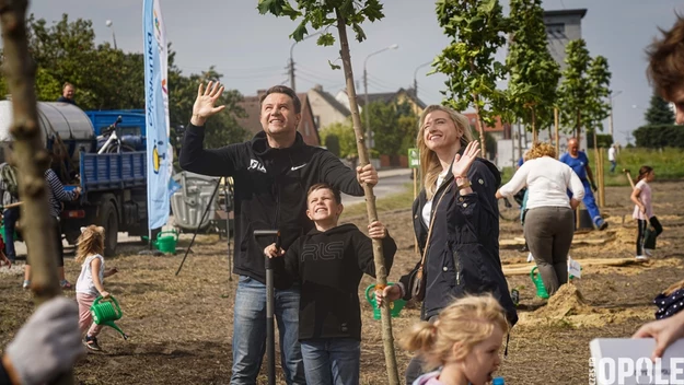 Grupa ludzi biorąca udział w sadzeniu drzew, rodzina w centrum uśmiecha się i macha, dziecko trzyma młode drzewko, w tle inni uczestnicy akcji oraz zielone sadzonki na otwartym terenie miejskim, widoczne pozytywne emocje i zaangażowanie społeczności.