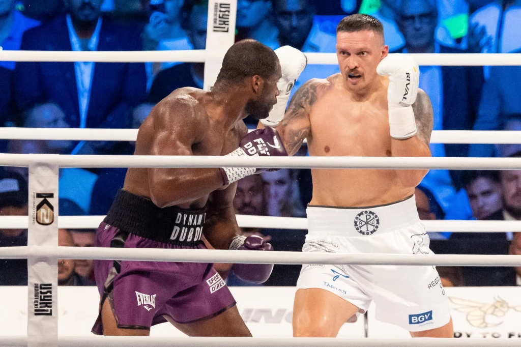 Ołeksandr Usyk tu w walce z Danielem Duboisem Dwóch bokserów walczących na ringu otoczonych przez publiczność, jeden z zawodników uderza, a drugi przyjmuje pozycję obronną, scena dynamiczna, tłem jest tłum widzów obserwujących pojedynek.