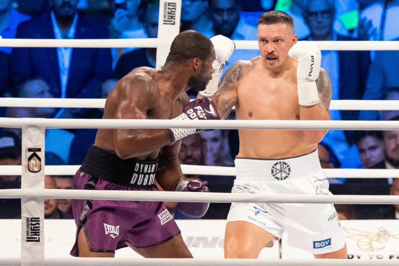 Dwóch bokserów walczących na ringu otoczonych przez publiczność, jeden z zawodników uderza, a drugi przyjmuje pozycję obronną, scena dynamiczna, tłem jest tłum widzów obserwujących pojedynek.