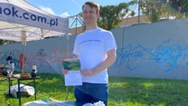 Mężczyzna stojący przy stoliku z ulotkami, książkami i materiałami promocyjnymi, w tle namiot oraz mur z graffiti, pogoda słoneczna, teren zielony.