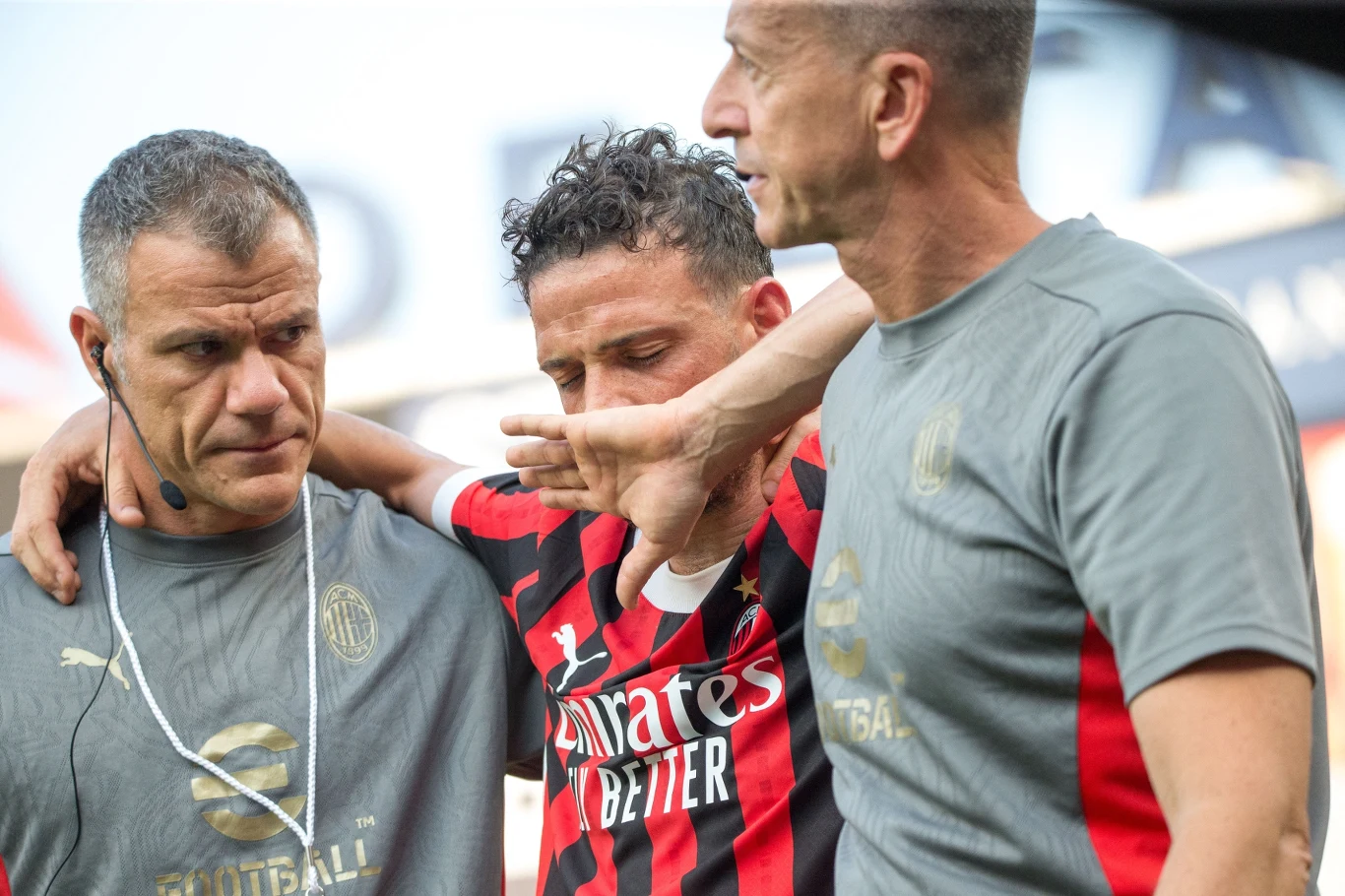 Piłkarz w koszulce w czerwono-czarne pasy, wspierany przez dwóch członków sztabu medycznego klubu AC Milan, wygląda na kontuzjowanego lub rozemocjonowanego, opierając się na ramionach obu osób.