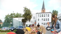Dzieci ustawione wzdłuż długiego stołu zajmują się sadzeniem roślin do małych doniczek, a dorosła kobieta w długiej spódnicy instruuje je jak to robić; w tle widoczny budynek szkolny, samochody i fragment miasta.