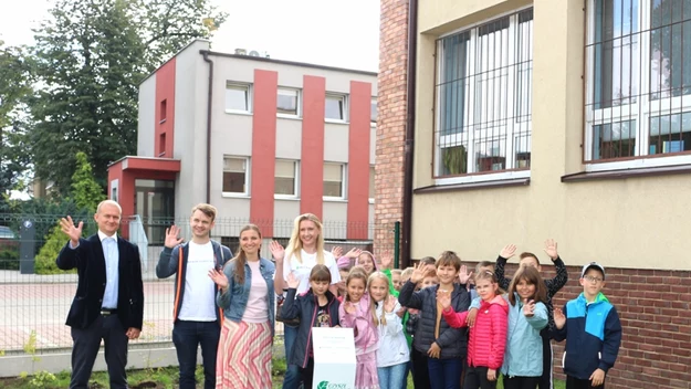 Grupa dzieci i osób dorosłych stoi przed budynkiem szkoły, wszyscy uśmiechają się i machają do obiektywu, na środku widoczna tablica lub plakat z logo Zielonej Szkoły.