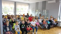 Grupa dzieci w wieku szkolnym siedząca na krzesłach w jasno oświetlonym pomieszczeniu z dużymi oknami, wśród nich kilku dorosłych opiekunów, wszyscy zwróceni w stronę niewidocznego prowadzącego.