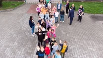 Grupa dzieci i dorosłych ustawiona na placu w kształt litery, większość dzieci macha do obiektywu, dorośli stoją z tyłu, wszyscy na tle brukowanego placu przed budynkiem.