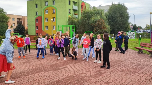 Grupa dzieci bawi się wspólnie na świeżym powietrzu w pobliżu kolorowego bloku mieszkalnego, nadzorowana przez dorosłą kobietę podnoszącą rękę, w tle widać plac zabaw, drzewa i ławki.