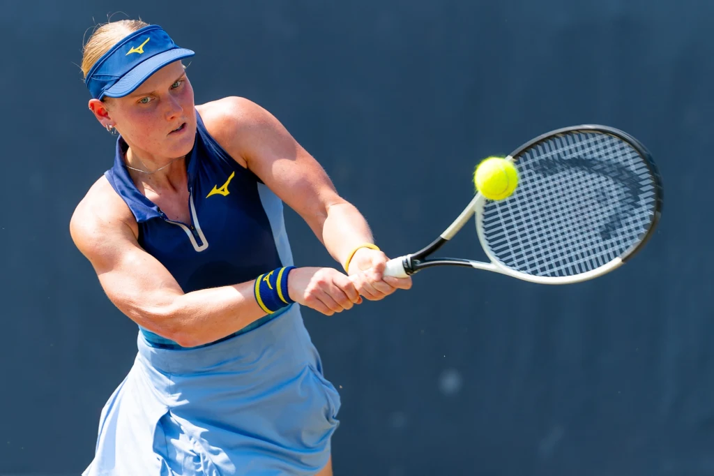 Suzan Lamens podczas Wimbledonu 2025 Tenisistka w niebieskiej sportowej sukience i czapce, skoncentrowana podczas uderzania piłki żółtą rakietą tenisową na korcie.