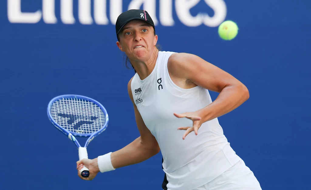 Iga Świątek w trakcie US Open 2025 Tenisistka w białym stroju podczas dynamicznego zamachu rakietą, skupiona na żółtej piłce tenisowej na tle niebieskiego kortu.