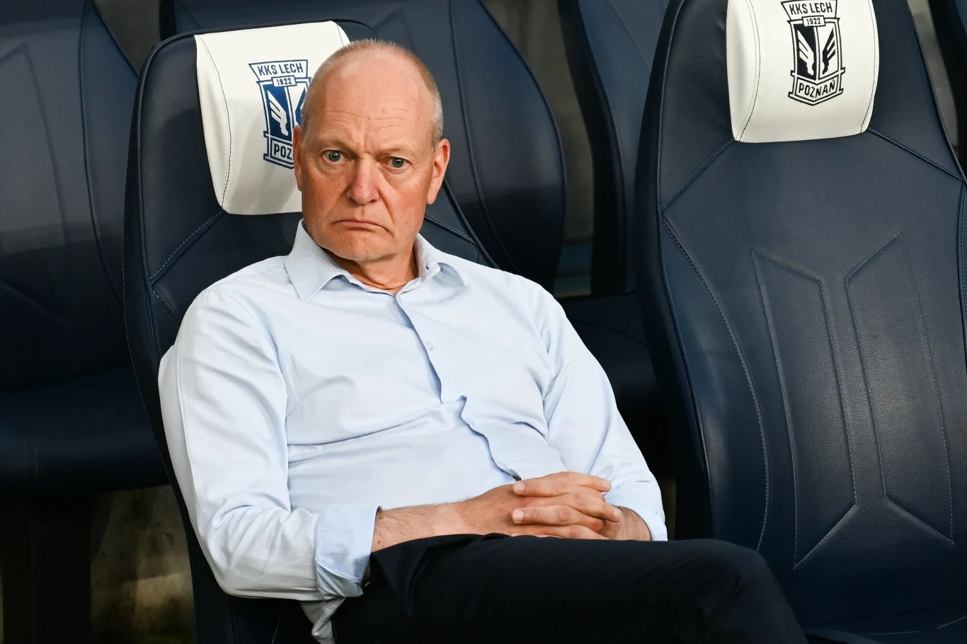 Starszy mężczyzna o poważnym wyrazie twarzy siedzi na stadionowej ławce trenerskiej, ubrany w jasnoniebieską koszulę, w tle widoczne są logo klubu piłkarskiego Lech Poznań na oparciach foteli.