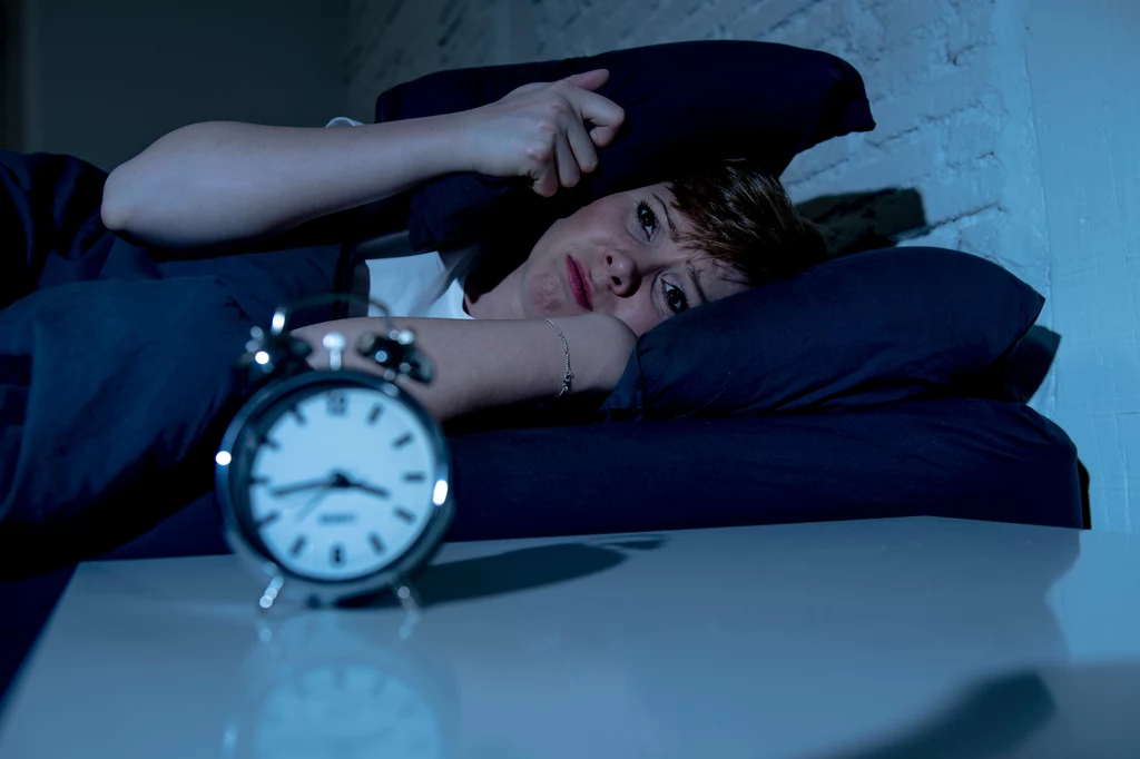 Problemy ze snem jesienią są częste. Można jednak temu przeciwdziałać Kobieta leżąca w łóżku, zakrywająca głowę poduszką z niezadowoloną lub zmartwioną miną, na pierwszym planie stoi klasyczny budzik wskazujący wczesną godzinę.