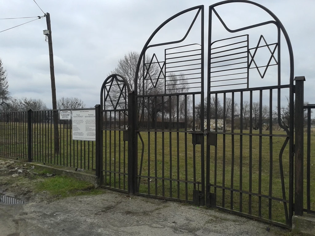 Metalowa brama z motywem gwiazdy Dawida, otoczona ogrodzeniem, prowadzi na teren zielonego, ogrodzonego cmentarza żydowskiego, z tablicą informacyjną zamontowaną na płocie po lewej stronie, w tle widoczne nagrobki i bezlistne drzewa oraz pochmurne niebo.