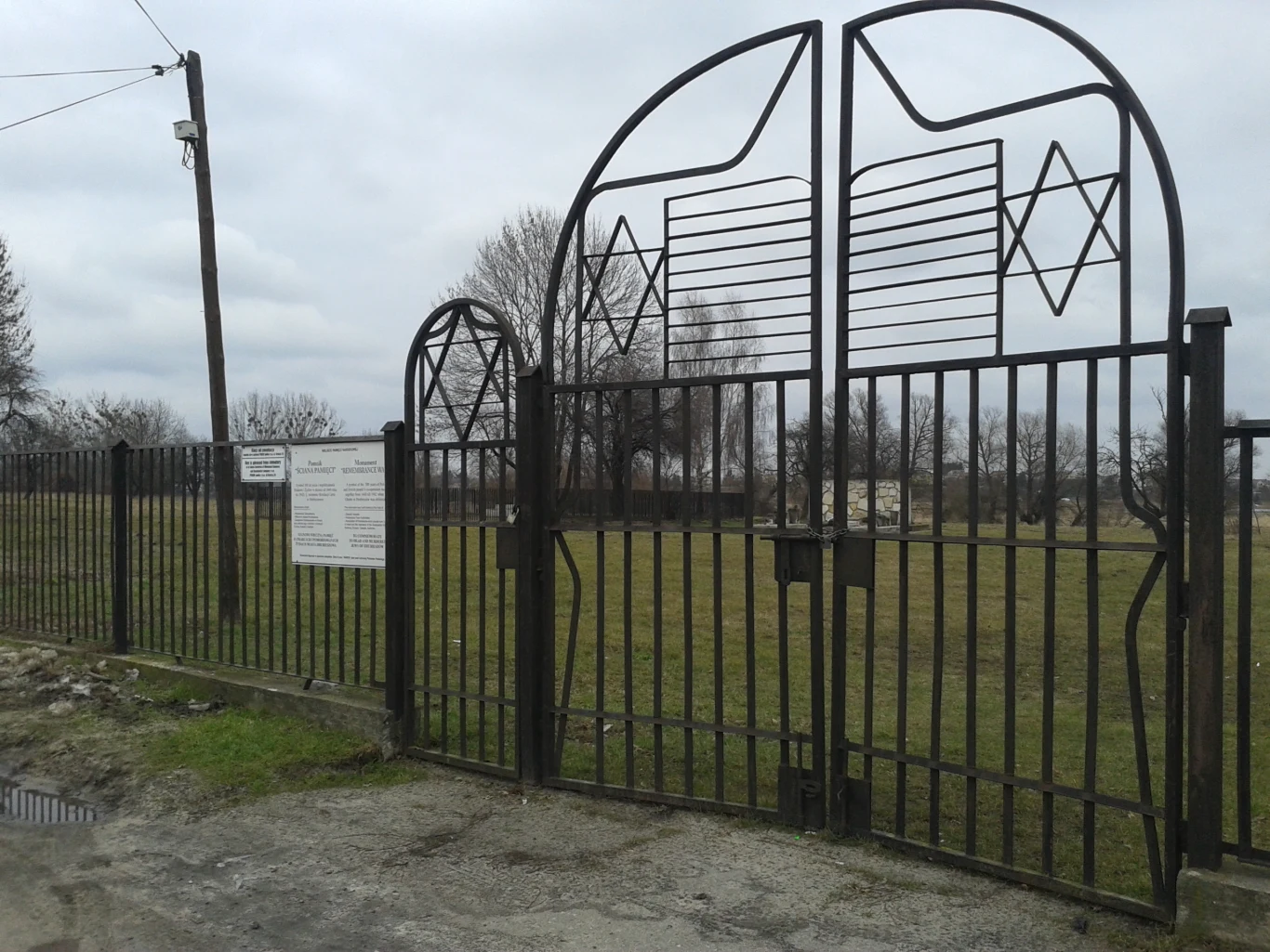 Metalowa brama z motywem gwiazdy Dawida, otoczona ogrodzeniem, prowadzi na teren zielonego, ogrodzonego cmentarza żydowskiego, z tablicą informacyjną zamontowaną na płocie po lewej stronie, w tle widoczne nagrobki i bezlistne drzewa oraz pochmurne niebo.