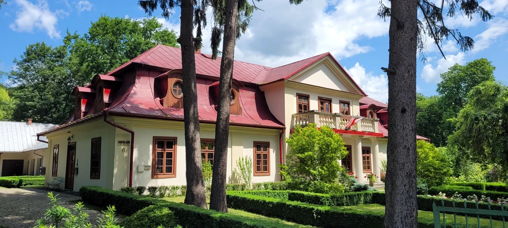 Dworek z czerwonym dachem i ozdobną balustradą, otoczony zadbanym ogrodem z geometrycznymi żywopłotami, wśród wysokich drzew i zieleni.