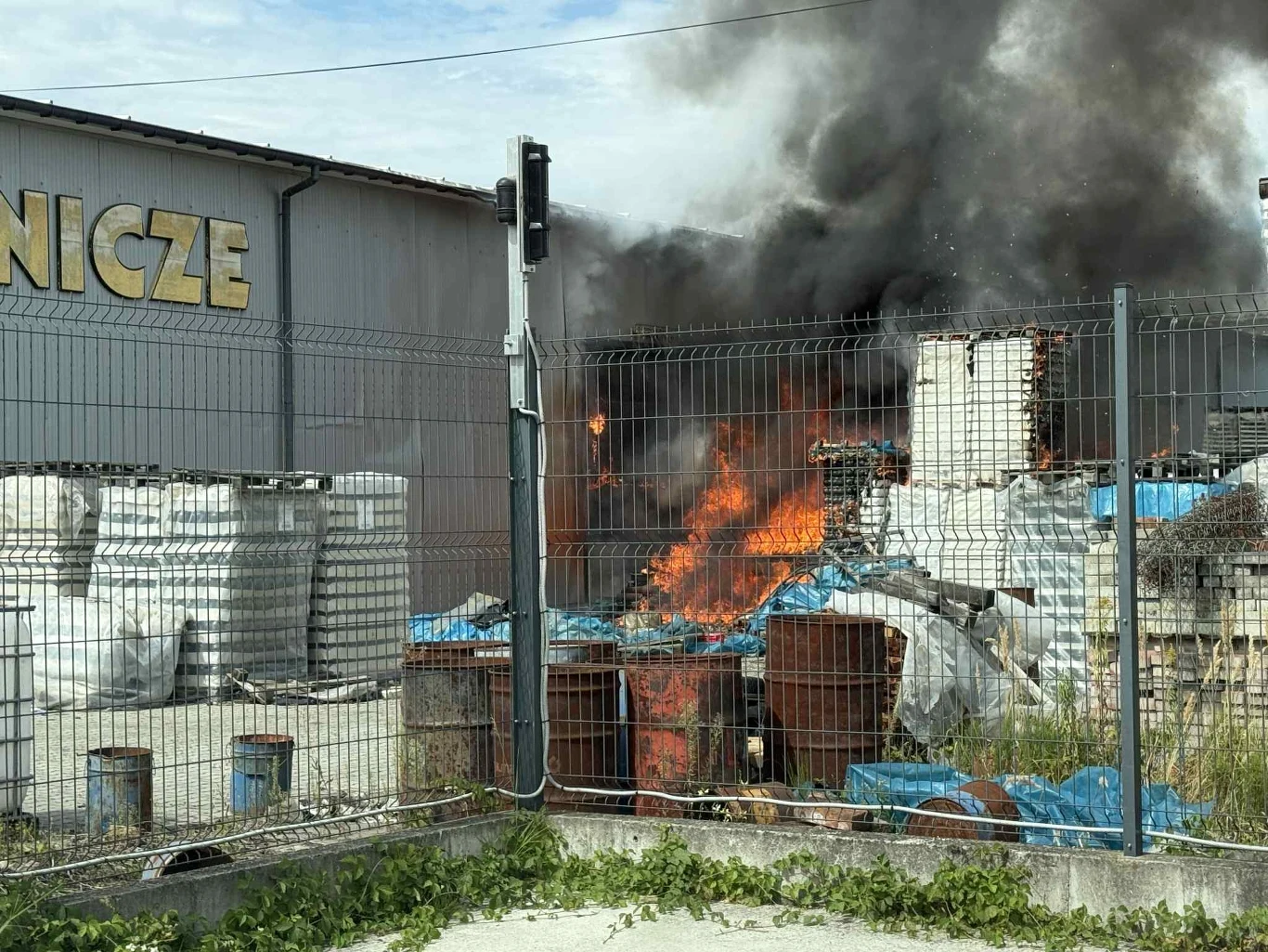 Tarnów. Pożar w hurtowni zniczy. Na miejscu 25 zastępów Intensywny pożar na terenie przemysłowym, płomienie trawiące materiały składowane na zewnątrz magazynu, nad płonącymi beczkami i materiałami unosi się gęsty, czarny dym.