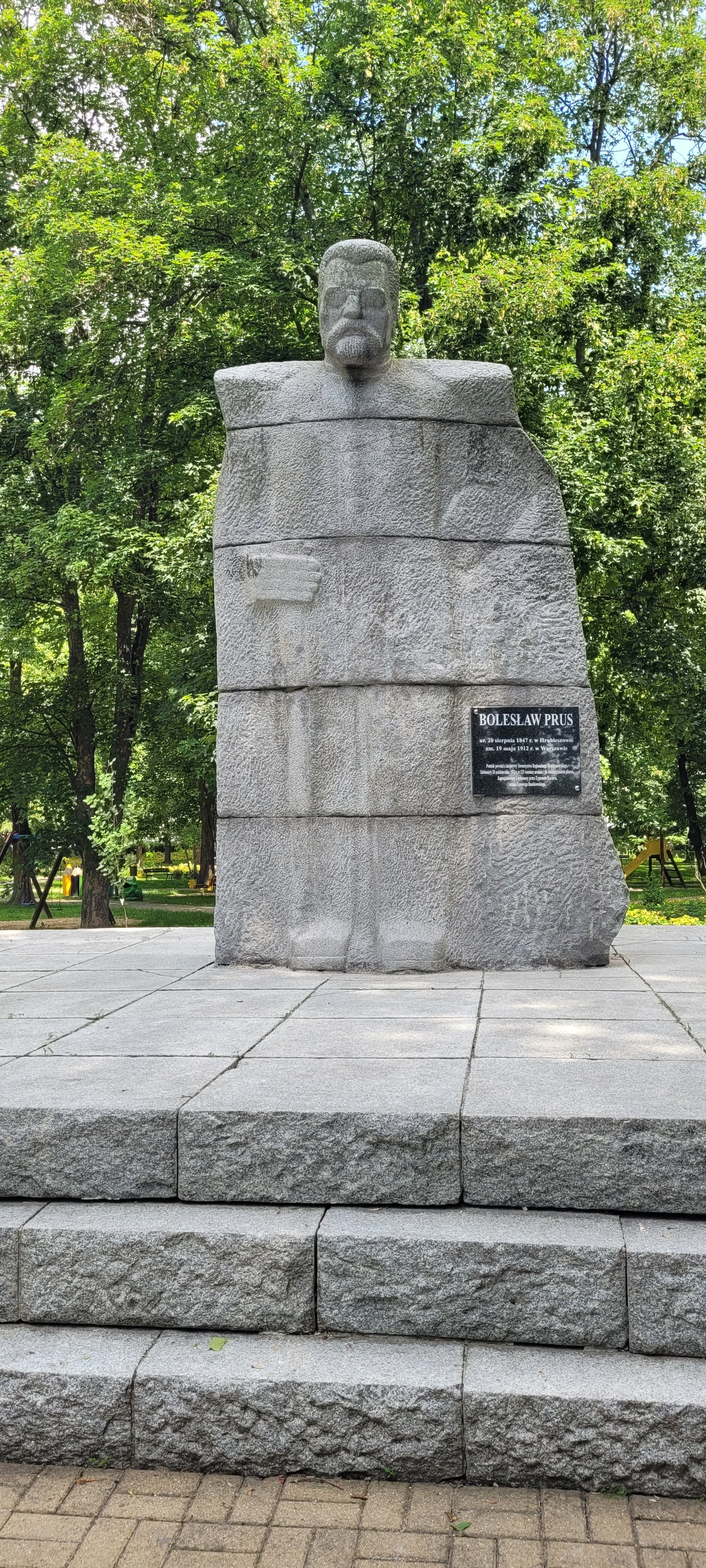 Kamienny pomnik z wizerunkiem popiersia mężczyzny, ustawiony na schodkowym podeście, wokół zielone drzewa parku, na froncie monumentu znajduje się tablica pamiątkowa.