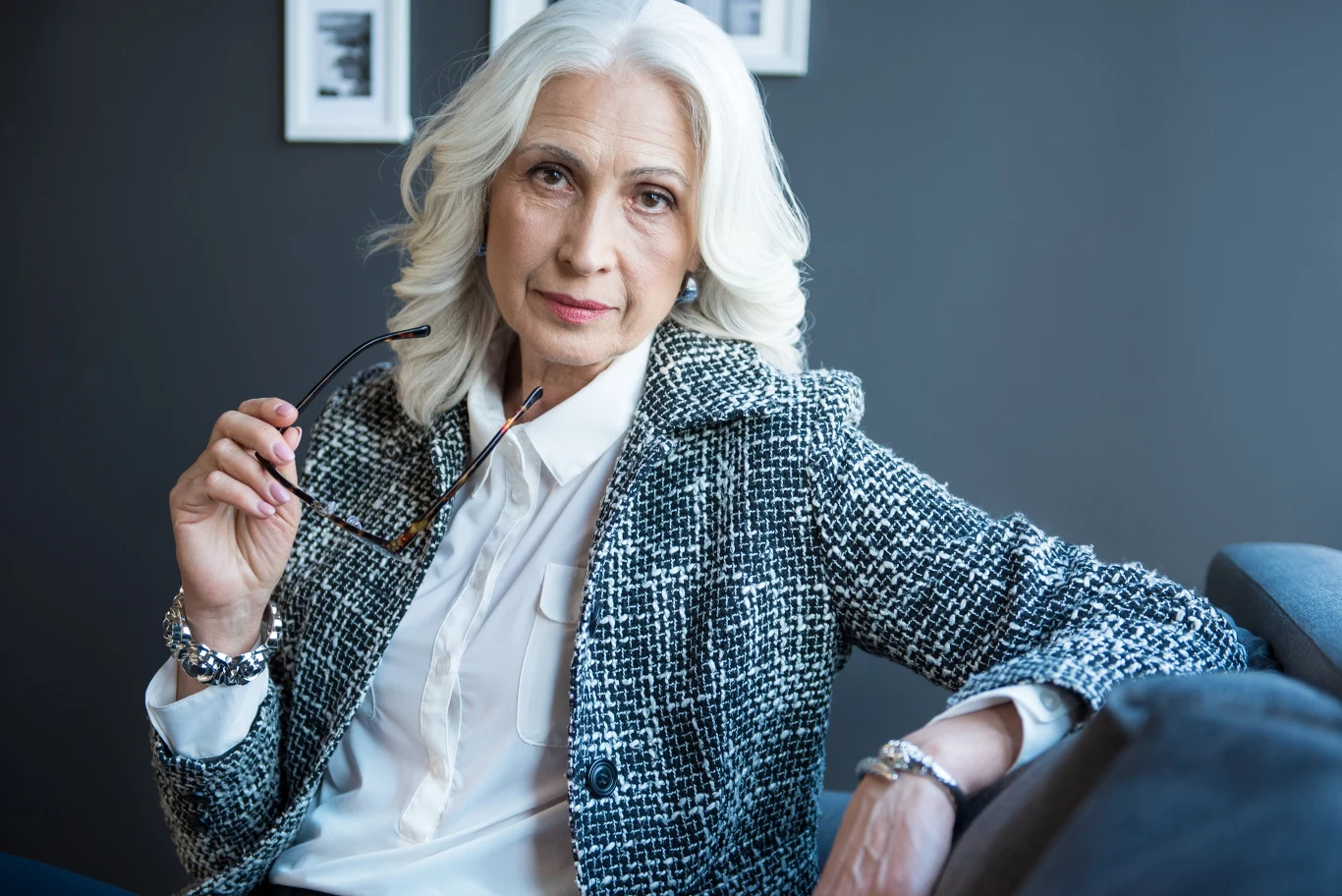 Wróżka Diana ułożyła tarota dla wszystkich znaków. Sprawdź, co się wydarzy w piątek Starsza elegancka kobieta z siwymi włosami w białej koszuli i stylowej marynarce trzyma okulary w dłoni i siedzi na kanapie, patrząc prosto w obiektyw na tle ciemnej ściany z dwoma obrazami.