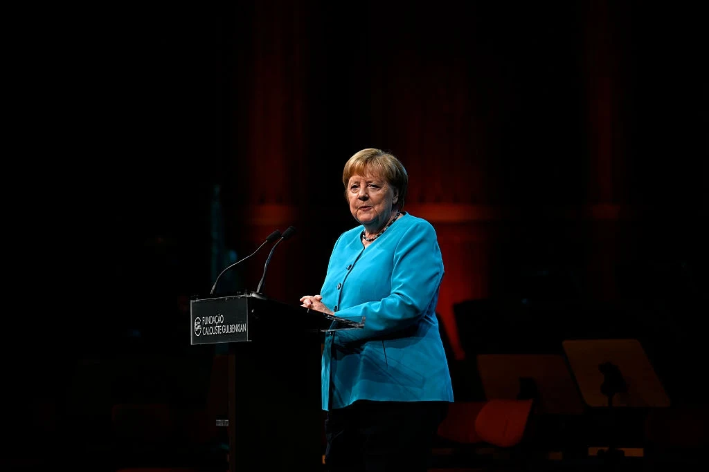 10 lat po słowach Angeli Merkel. Co się zmieniło? Kobieta w niebieskiej marynarce stoi za mównicą na scenie z mikrofonem, w tle widoczne ciemne otoczenie oraz rozmyte elementy sali.