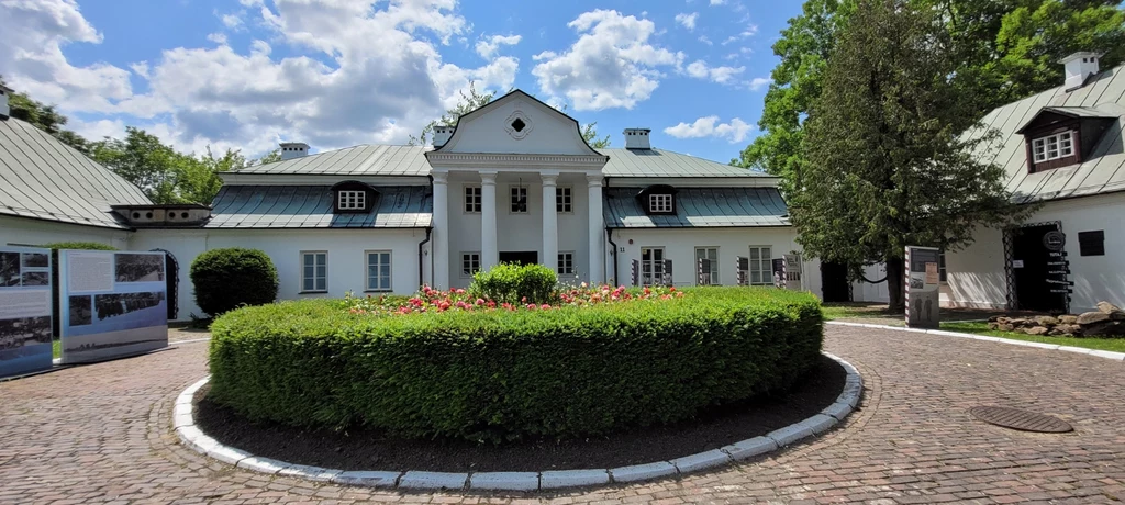 Klasyczny dwór z białą elewacją i czterokolumnowym portykiem, w centrum podjazdu zielone, zadbane rondo z krzewami i kwiatami, otoczenie tworzą niskie budynki oraz stare drzewa, po lewej stronie widoczne tablice informacyjne.