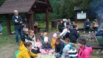 Grupa dzieci i dorosłych siedzących na kocu oraz przy ognisku w otoczeniu drewnianych altanek i lasu, atmosfera wspólnego spędzania czasu i integracji na łonie natury.
