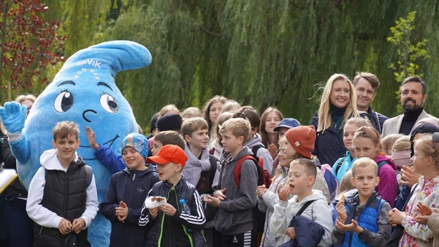 Grupa dzieci i dorosłych zebrana na świeżym powietrzu, większość dzieci w kurtkach i sportowych ubraniach, w centrum postać maskotki w niebieskim kostiumie graficznym z uśmiechem i wielkimi oczami, otoczenie zielonych drzew i radosny nastrój.
