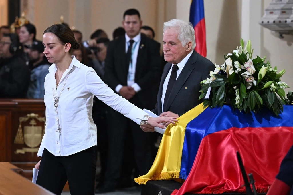 Miguel Uribe Londono zamierza ubiegać się o fotel prezydenta Kolumbii Trumna przykryta narodową flagą otoczona kwiatami, dwie osoby w żałobie oddające hołd zmarłemu, w tle widoczni żałobnicy oraz fotografowie dokumentujący wydarzenie.