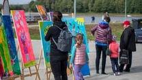 Kolorowe obrazy na sztalugach prezentowane na plenerowej wystawie, ludzie oglądający prace, wśród nich dzieci i dorośli spacerujący w pobliżu jeziora