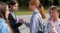 Grupa młodych dziewczyn na świeżym powietrzu, dwie z nich ściskają sobie dłonie w przyjacielskim geście, obok stoją pozostałe dziewczyny ubrane w sportowe bluzy, jedna z nich ma założone rękawiczki ochronne.